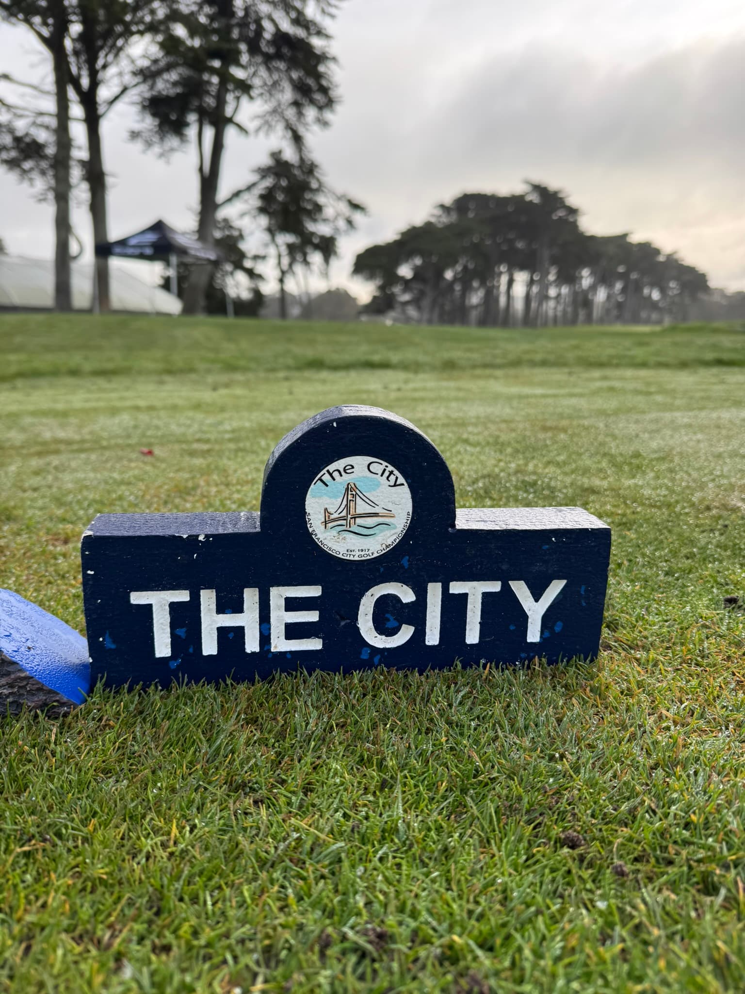 A weathered “The City” tee marker on the first tee, fog in the cypress beyond